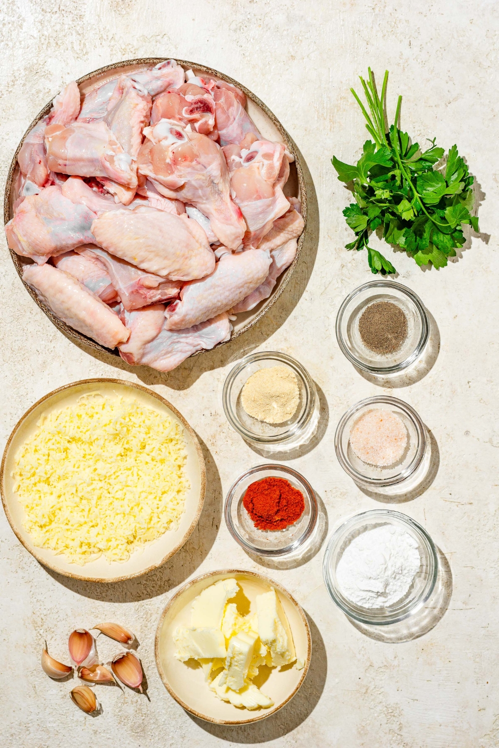 An overhead shot of several bowls in various sizes containing ingredients to make copycat Wingstop garlic parmesan chicken wings including chicken wings, parsley, grated parmesan cheese, butter, garlic, and seasonings.