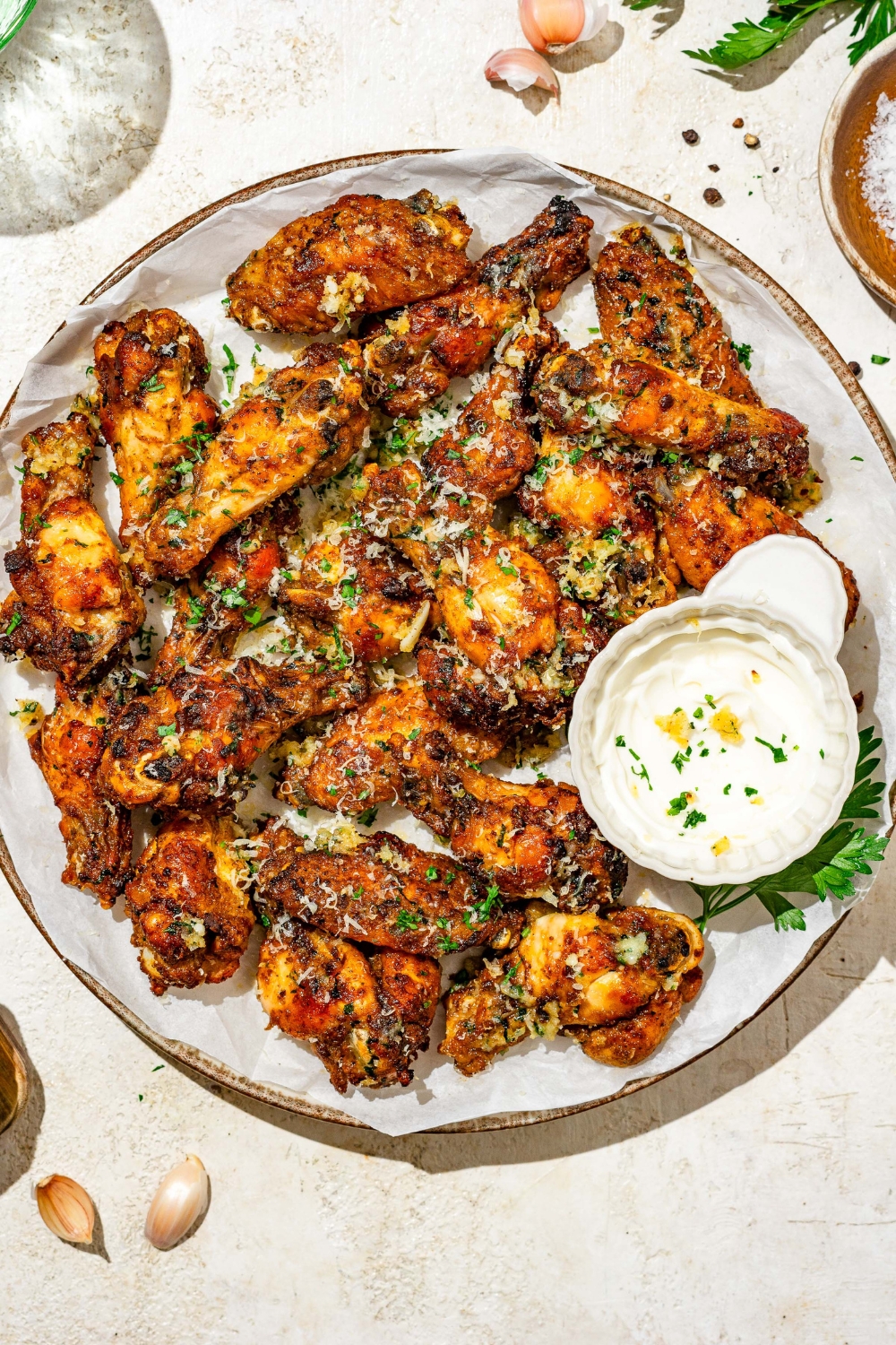 A plate of copycat Wingstop garlic parmesan chicken wings garnished with grated cheese and fresh parsley. There is a bowl of sauce on the plate. The plate is on a tan counter with small bowls of seasonings.