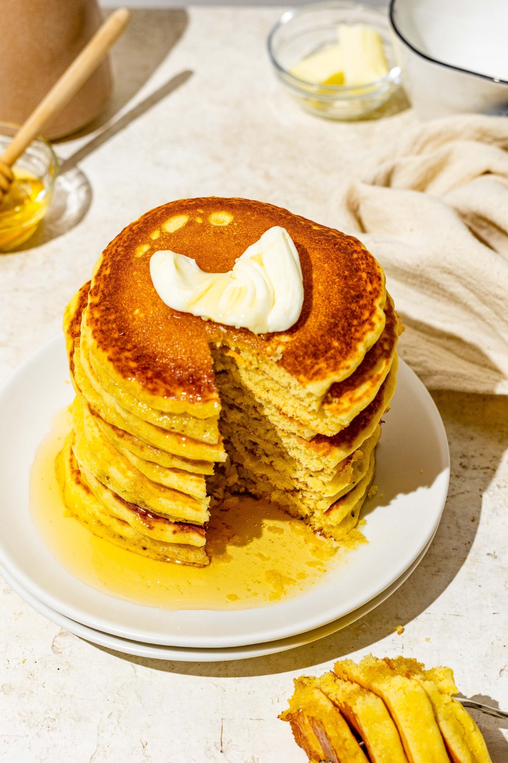 A white plate with a stack of cornmeal pancakes topped with butter and drizzled with syrup. A bite is taken from the stack. The plate is on a white counter with a fork with a bite of pancakes and white cloth napkin.