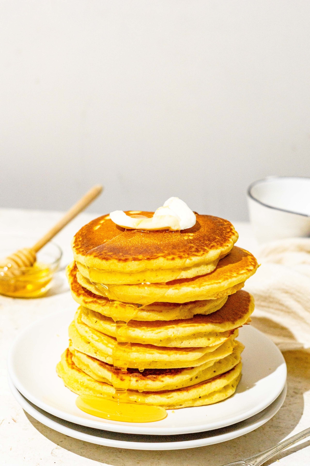 A white plate with a stack of cornmeal pancakes topped with butter and drizzled with syrup. The plate is on a white counter with a small bowl of syrup with a honey stirrer and white cloth napkin.