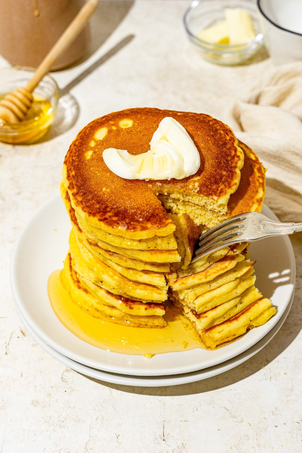 A white plate with a stack of cornmeal pancakes topped with butter and drizzled with syrup. A fork is taking a bite from the stack. The plate is on a white counter with a small bowl of syrup with a honey stirrer and white cloth napkin.