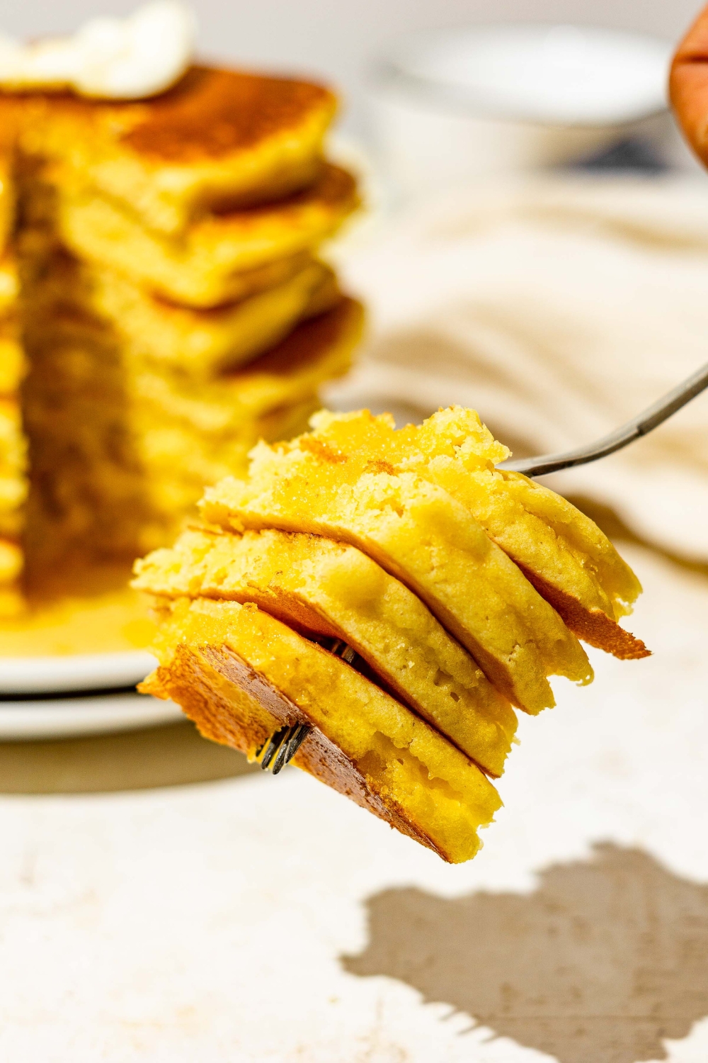 A close up of a fork with a bite from a stack of cornmeal pancakes. There is a white plate with pancakes on a white counter blurred in the background.