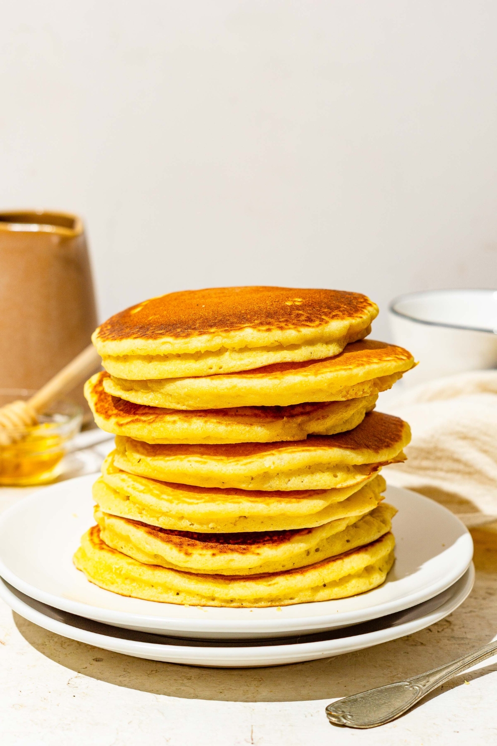 A white plate with a stack of cornmeal pancakes. The plate is on a white counter with a small bowl of syrup with a honey stirrer and white cloth napkin.