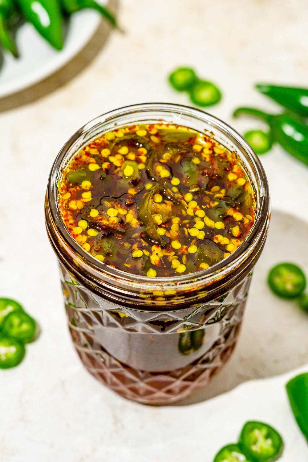A jar of cowboy candy candied jalapeños on a tan counter with sliced jalapeños.