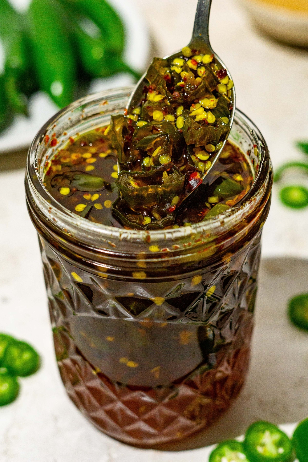 A jar of cowboy candy candied jalapeños on a tan counter with sliced jalapeños. There is spoon pouring jalapeños from the jar.