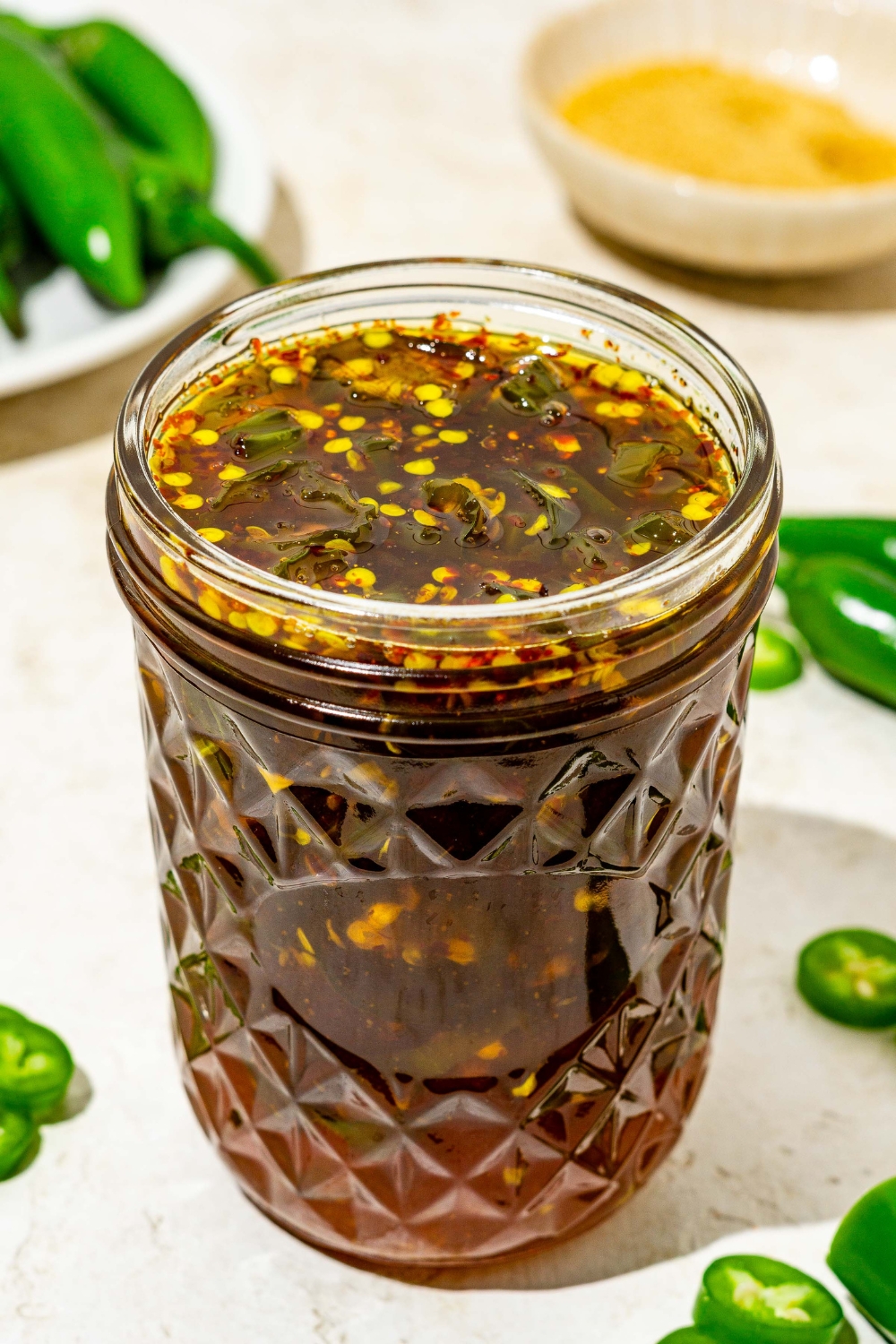 A jar of cowboy candy candied jalapeños on a tan counter with sliced jalapeños.