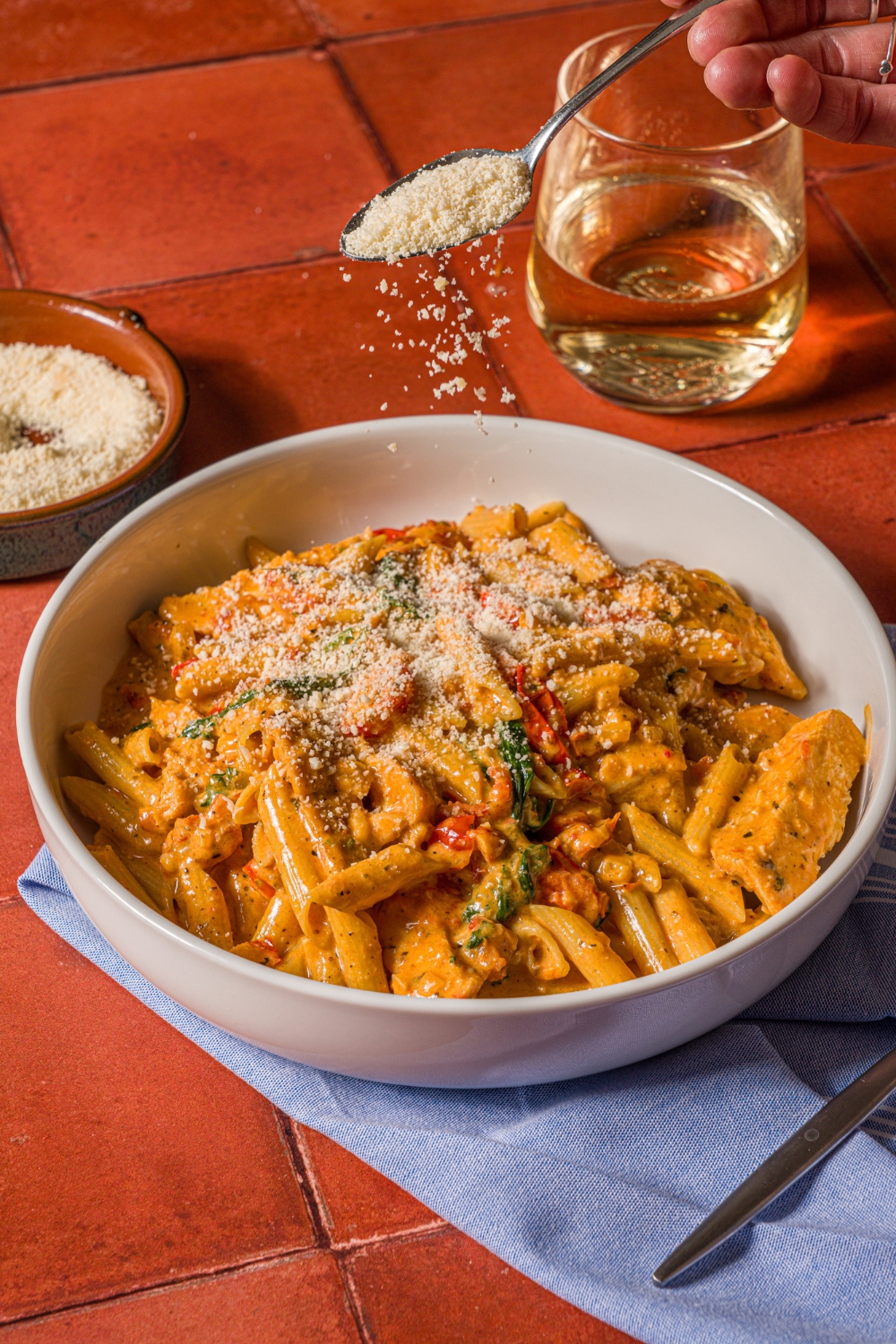 A white bowl with tuscan chicken pasta with a spoon of parmesan sprinkling over the bowl. The bowl is on a tiled counter with a blue cloth napkin, glass of white wine, and small bowl of parmesan.