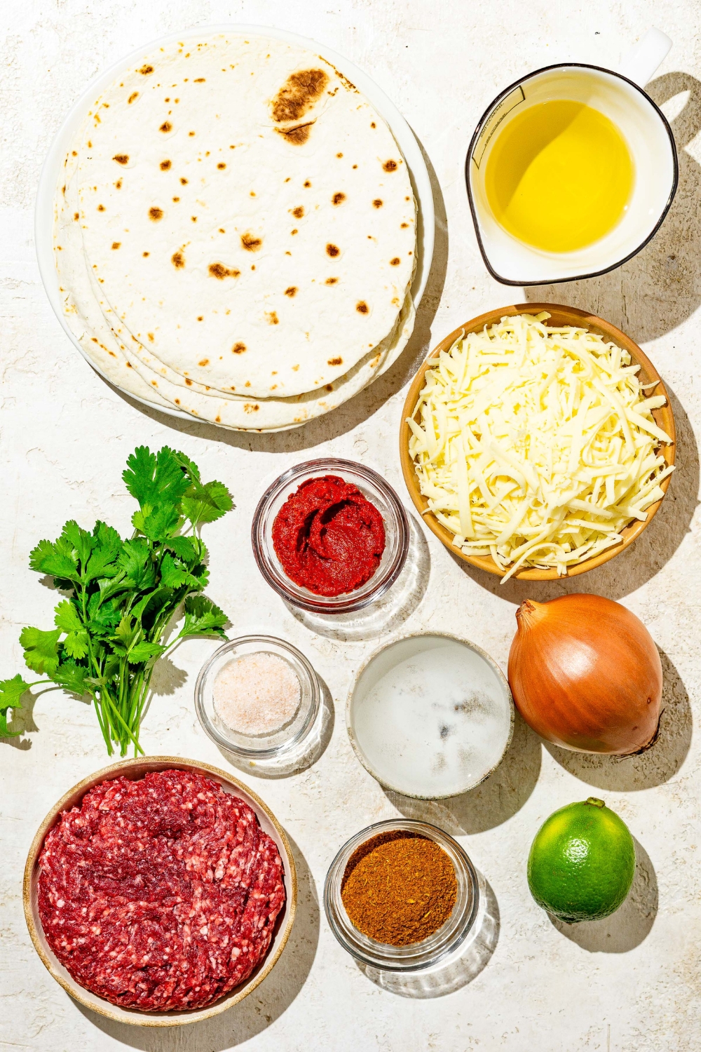 An overhead shot of several bowls in various sizes containing ingredients to make crispy beef tacos including tortillas, shredded mozzarella cheese, ground beef, onion, oil, lime, tomato paste, and seasonings.