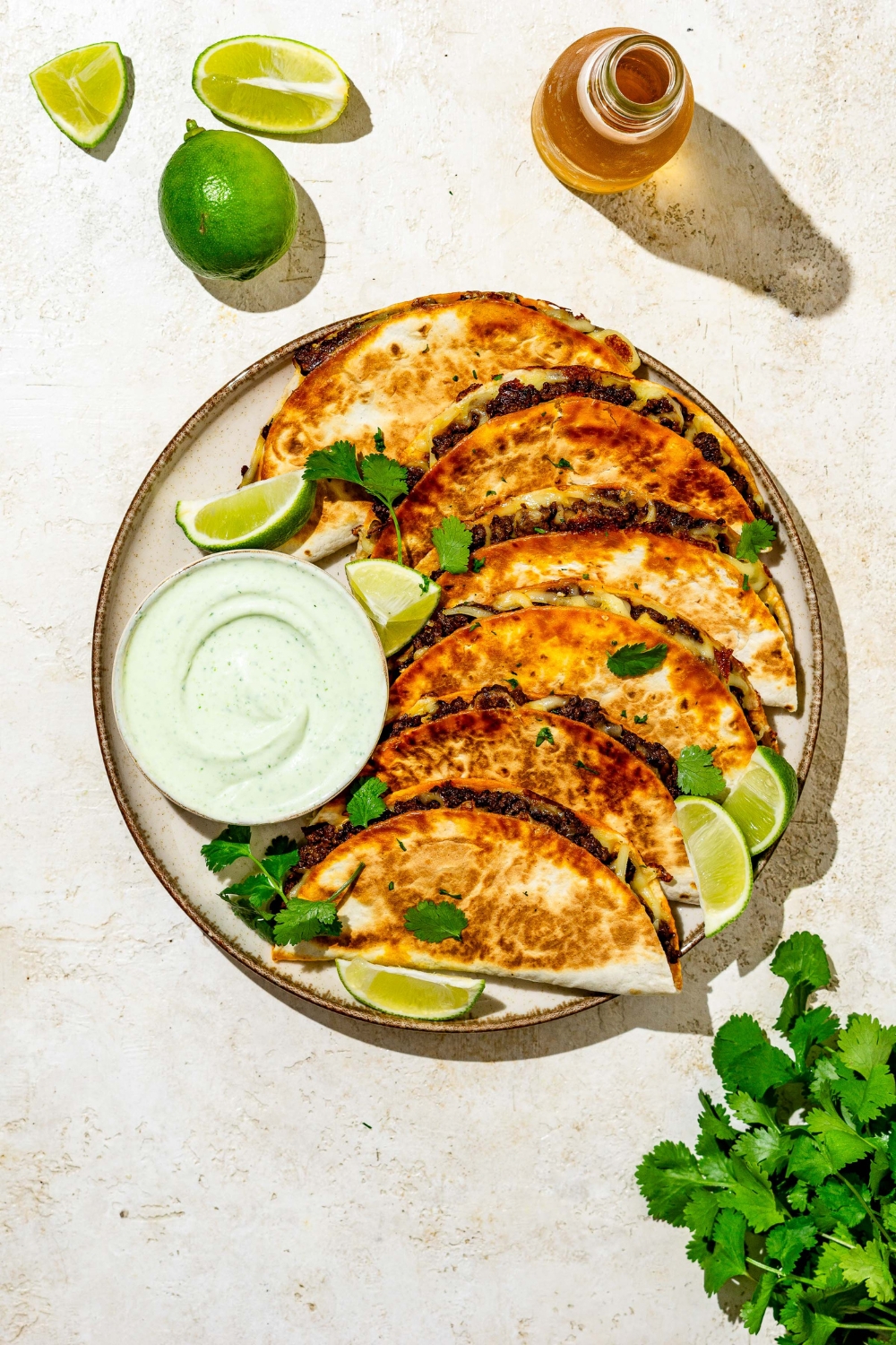 Crispy beef tacos layered on a plate garnished with cilantro and lime wedges and served with a side of sauce. The plate is on a tan counter with garnishes.