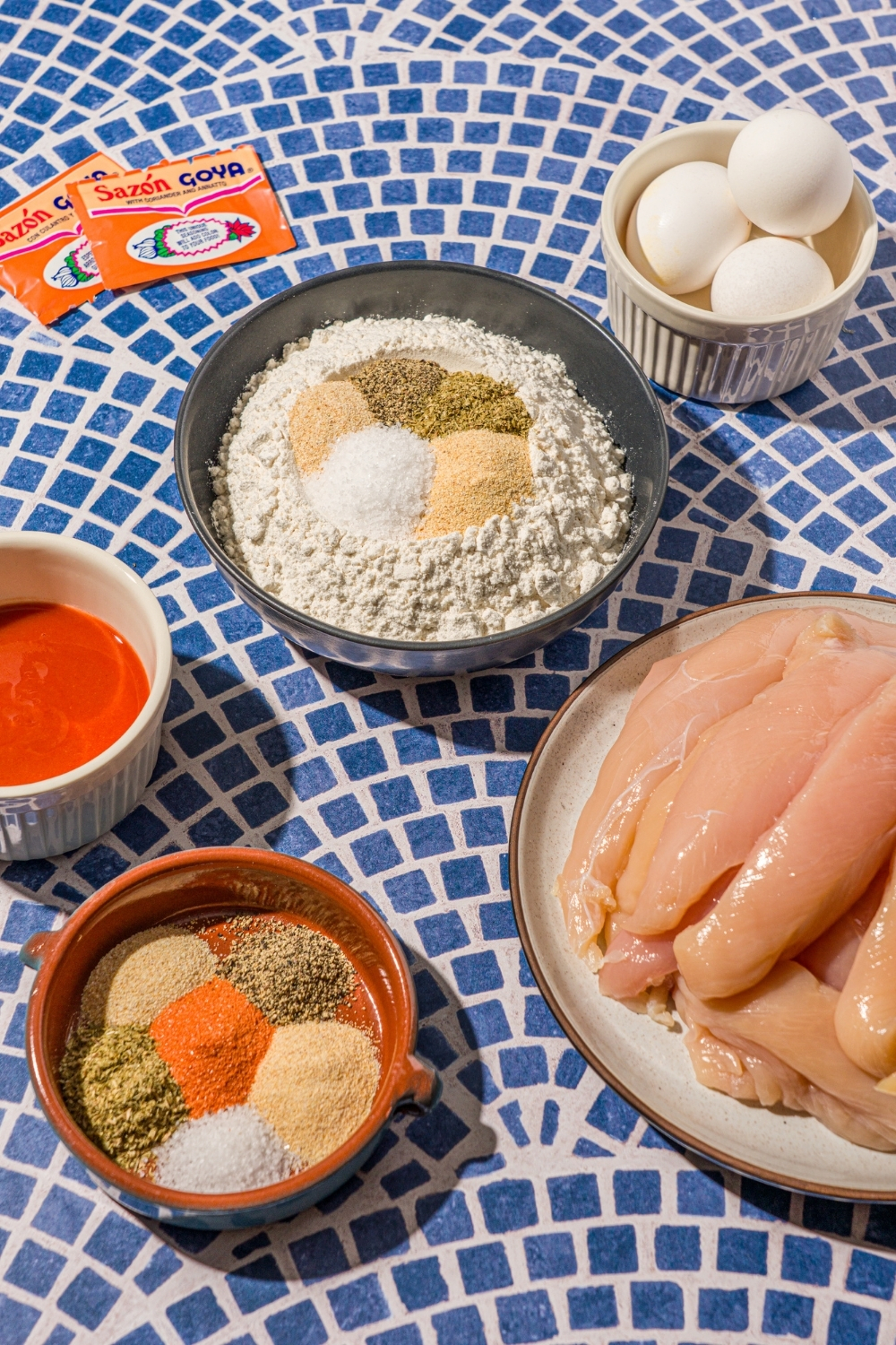 Several bowls in various sizes containing ingredients to make crispy chicken wraps including chicken tenders, hot sauce, eggs, and bowls of seasonings like flour, packets of sazon, garlic powder, onion powder, dried oregano, and more.