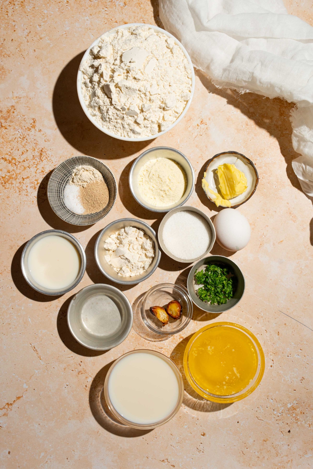 An overhead shot of several bowls in various sizes containing ingredients to make dinner rolls including flour, eggs, active yeast, sugar, butter, garlic, and chives.