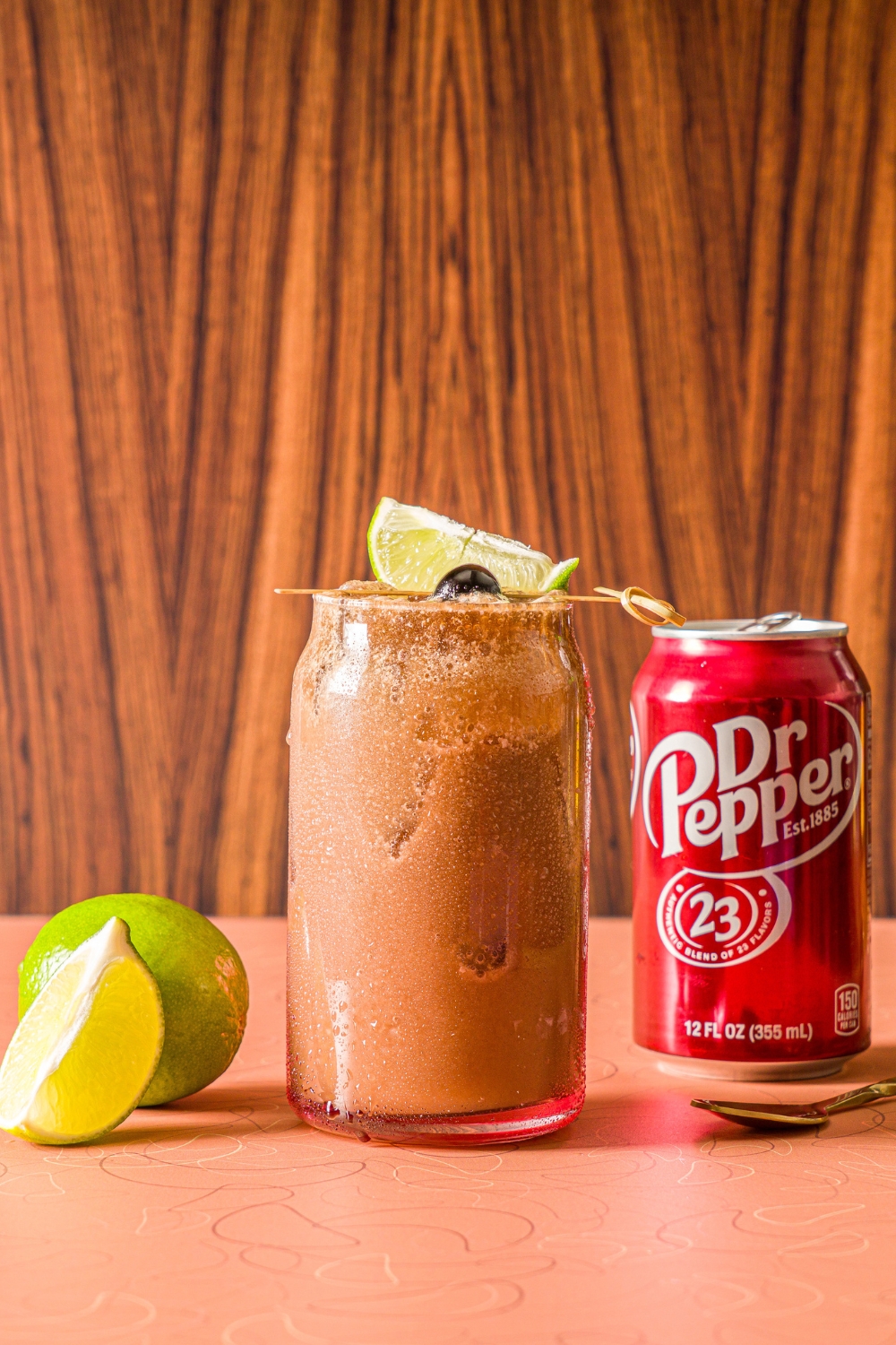 A glass of iced dirty soda garnished with a skewer of lime and maraschino cherry. The glass is on a counter iwth a can of Dr. Pepper and limes.