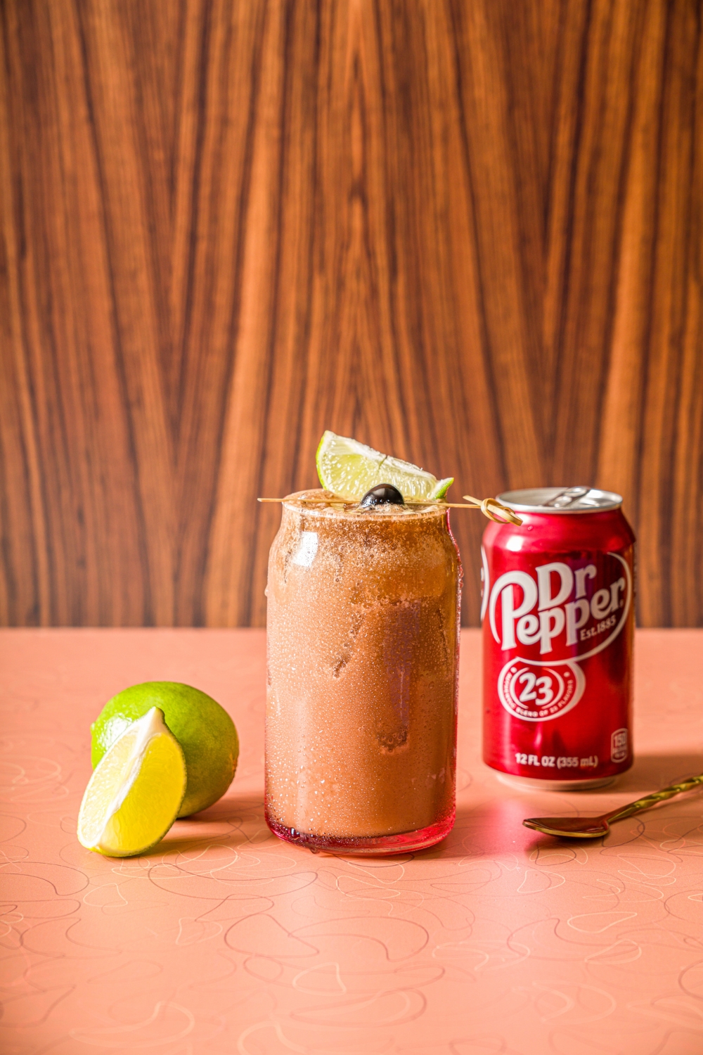 A glass of iced dirty soda garnished with a skewer of lime and maraschino cherry. The glass is on a counter iwth a can of Dr. Pepper and limes.