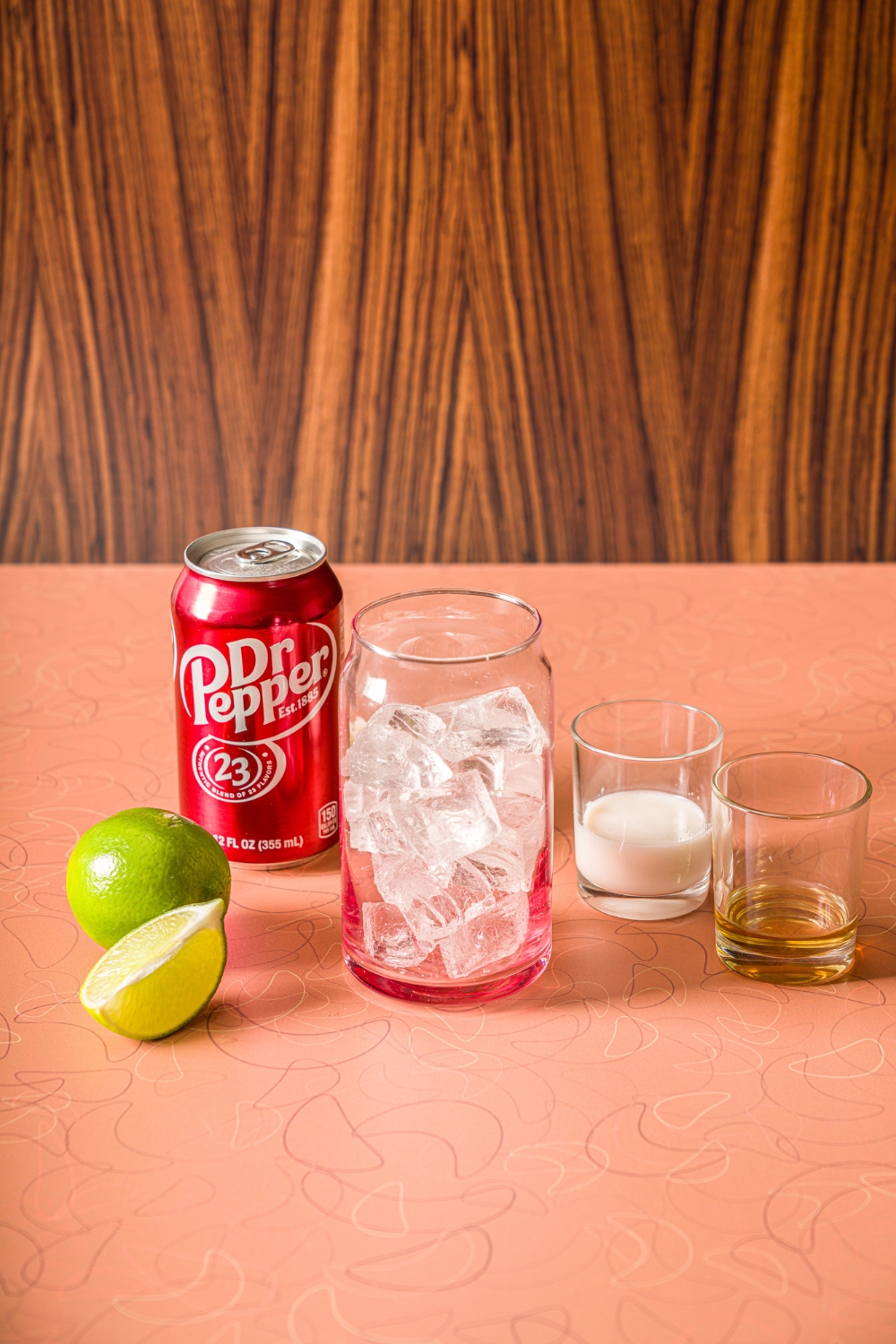 A counter with ingredients to make dirty soda including a glass of ice, a can of Dr. Pepper, a small glass of coconut creamer, a small glass of vanilla syrup, and limes.