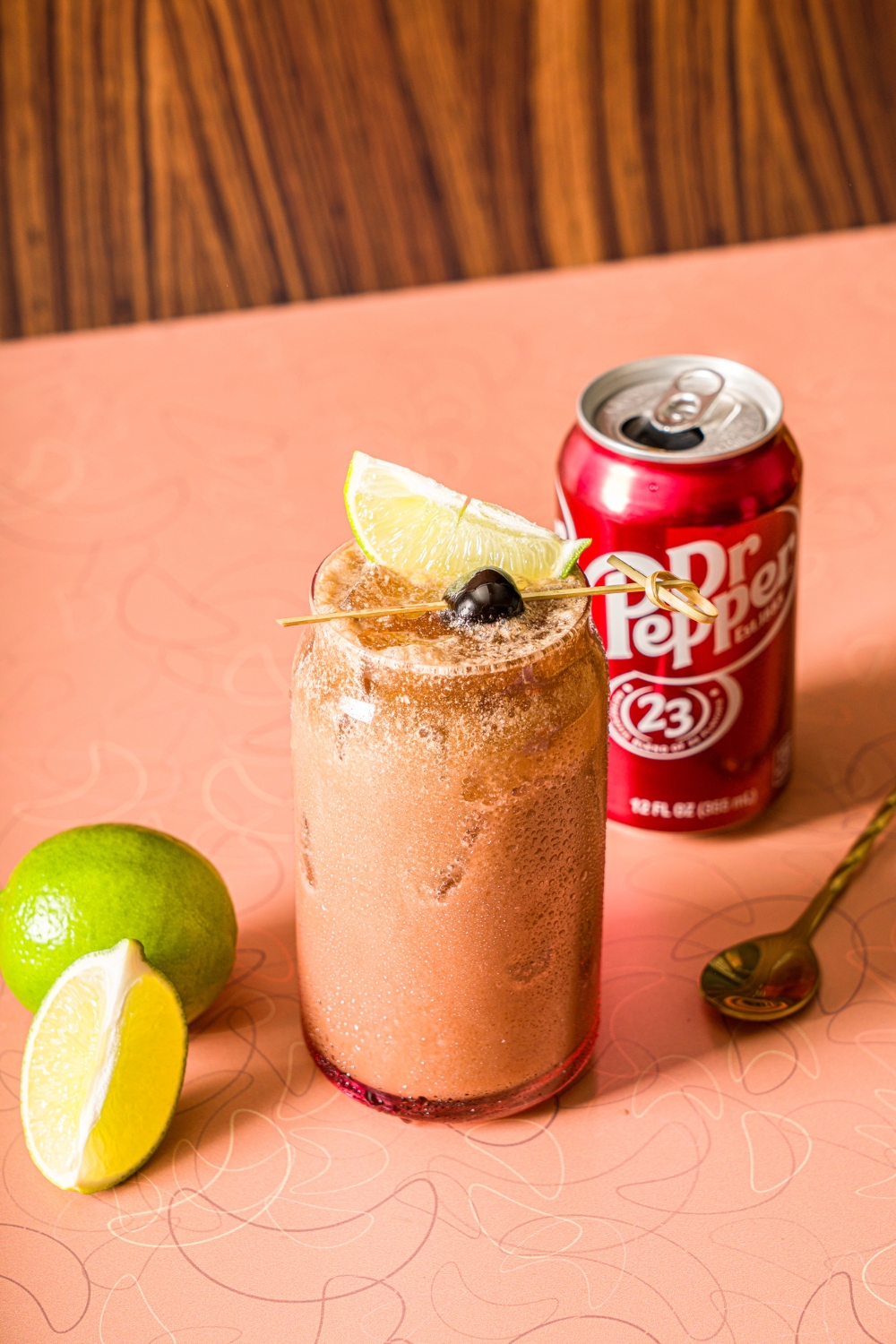 A glass of iced dirty soda garnished with a skewer of lime and maraschino cherry. The glass is on a counter iwth a can of Dr. Pepper and limes.