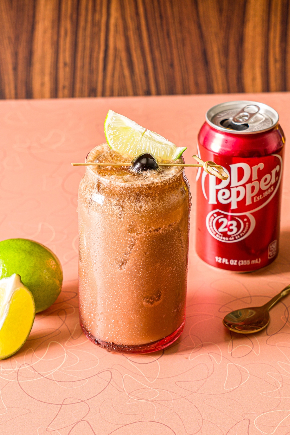 A glass of iced dirty soda garnished with a skewer of lime and maraschino cherry. The glass is on a counter iwth a can of Dr. Pepper and limes.