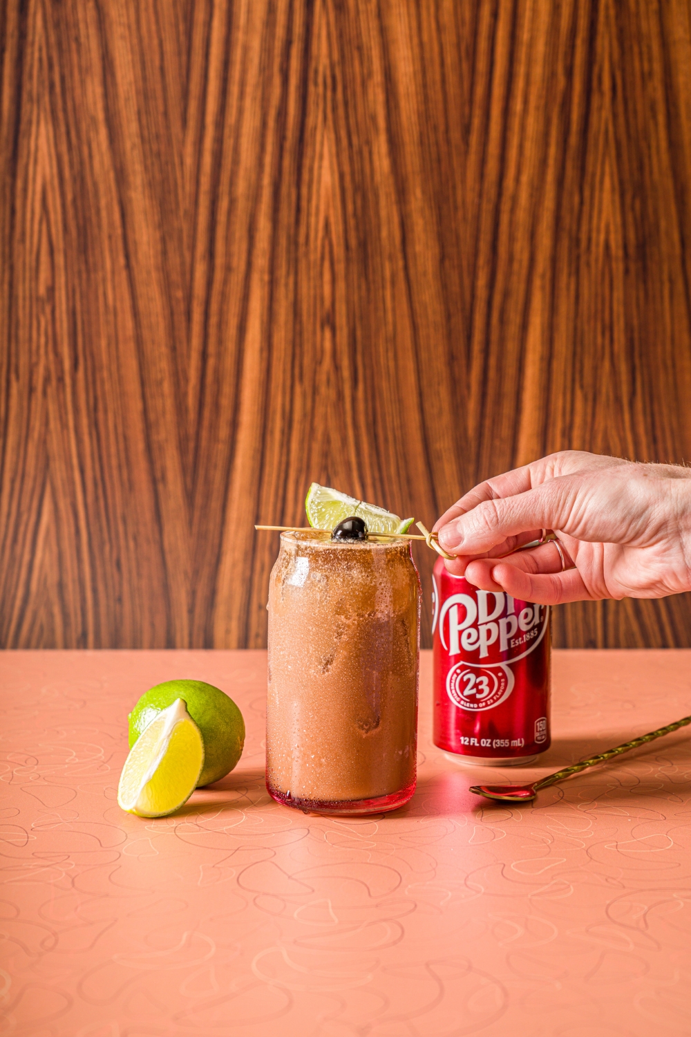 A glass of iced dirty soda garnished with a hand placing a skewer of lime and maraschino cherry on top. The glass is on a counter with a can of Dr. Pepper and limes.