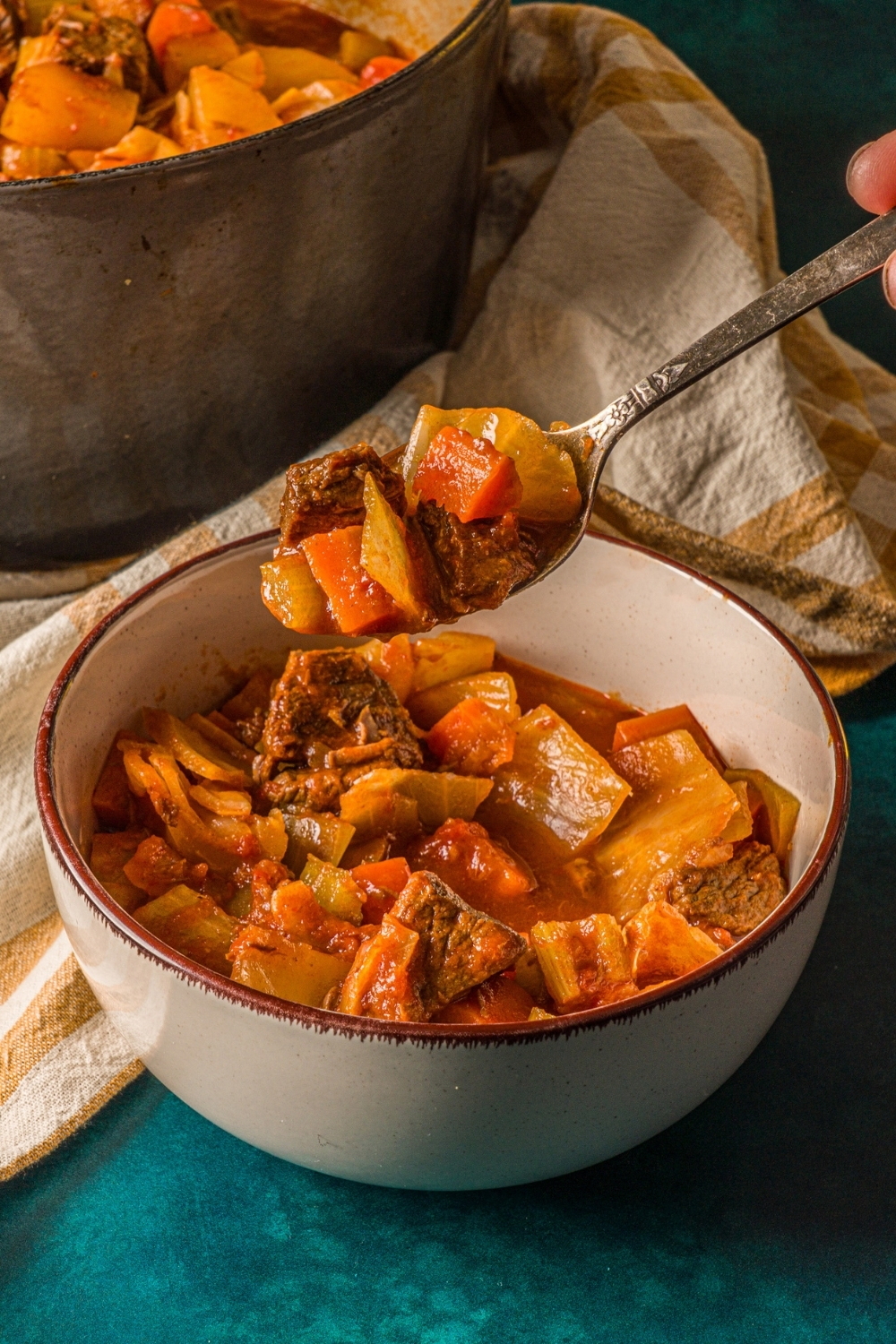A bowl of beef cabbage stew on a blue counter with a striped cloth napkin. There is a spoon with a bite of stew over the bowl.