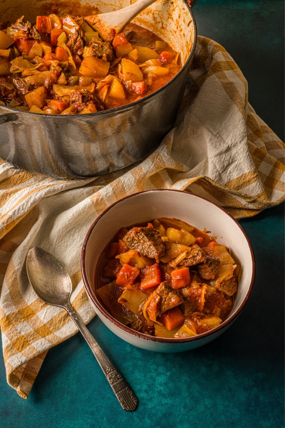 A bowl of beef cabbage stew on a blue counter with a spoon, striped napkin, and dutch oven with stew.
