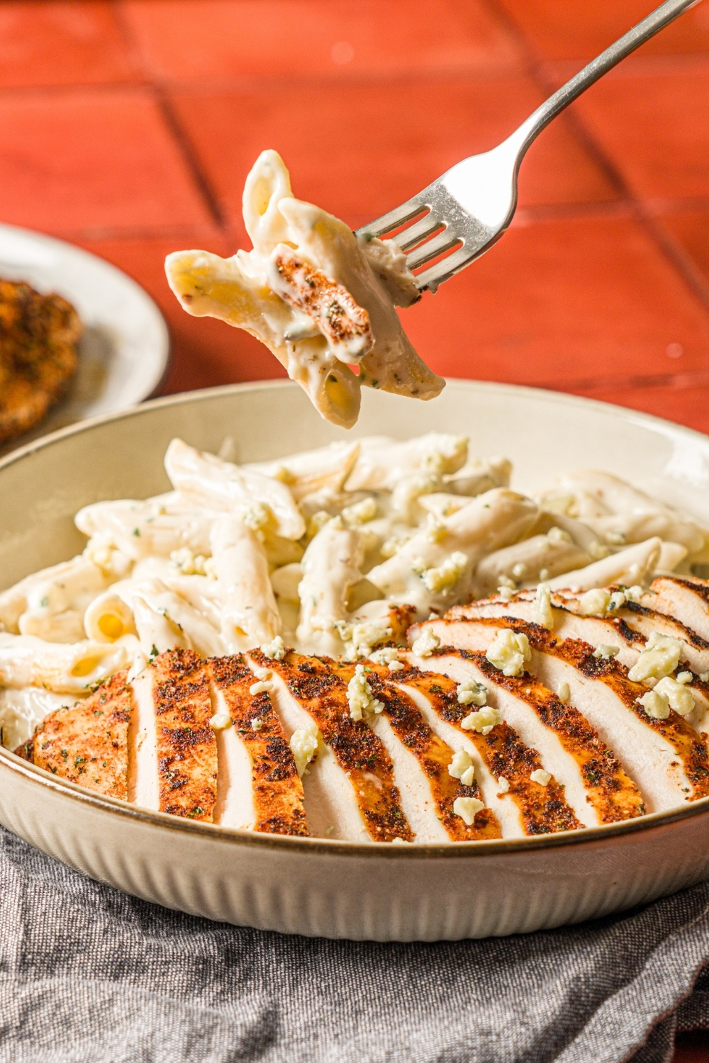 A bowl of blackened chicken alfredo with penne pasta garnished with bleu cheese. A fork is holding a bite of alfredo over the bowl. The bowl is on a red counter with a gray cloth napkin.