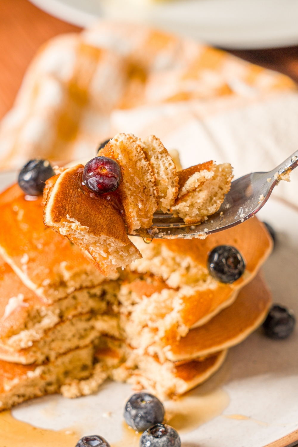 A close up of a fork with a bite of muffin mix pancakes. There is a plate of pancakes drizzled with syrup and served with blueberries in the background.