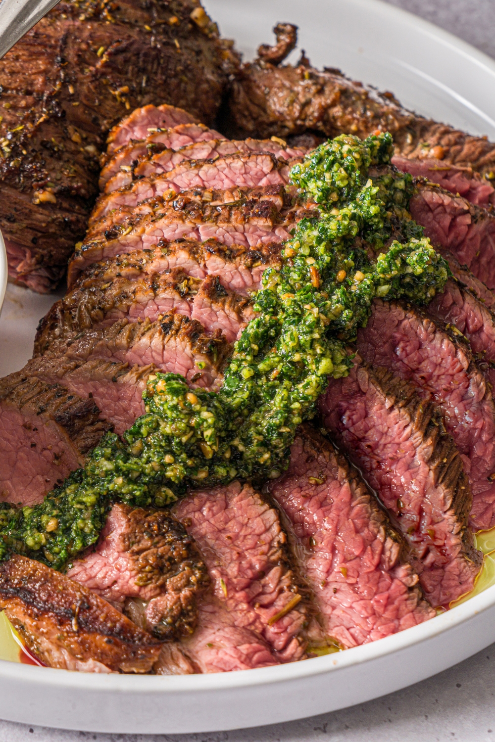 Sliced oven baked London broil on a white plate topped with pesto sauce. The plate is on a stone counter.