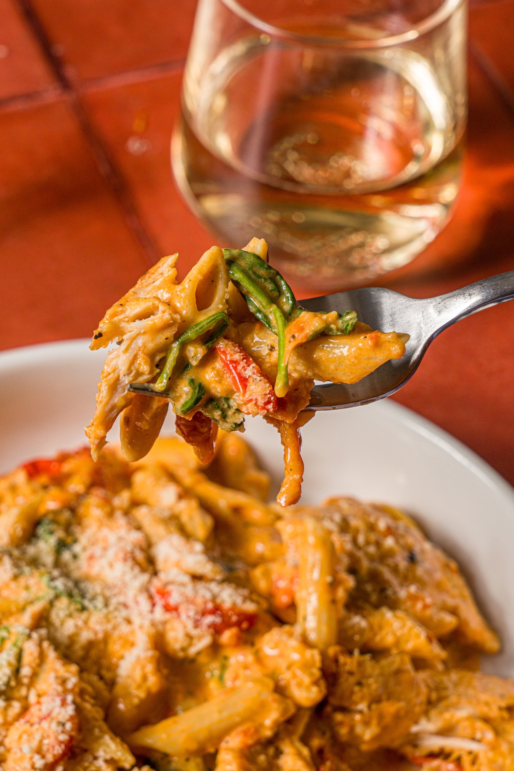 A close up of a fork with a bite of tuscan chicken pasta. The fork is over a white bowl of pasta. The bowl is on a tiled counter with a glass of white wine.
