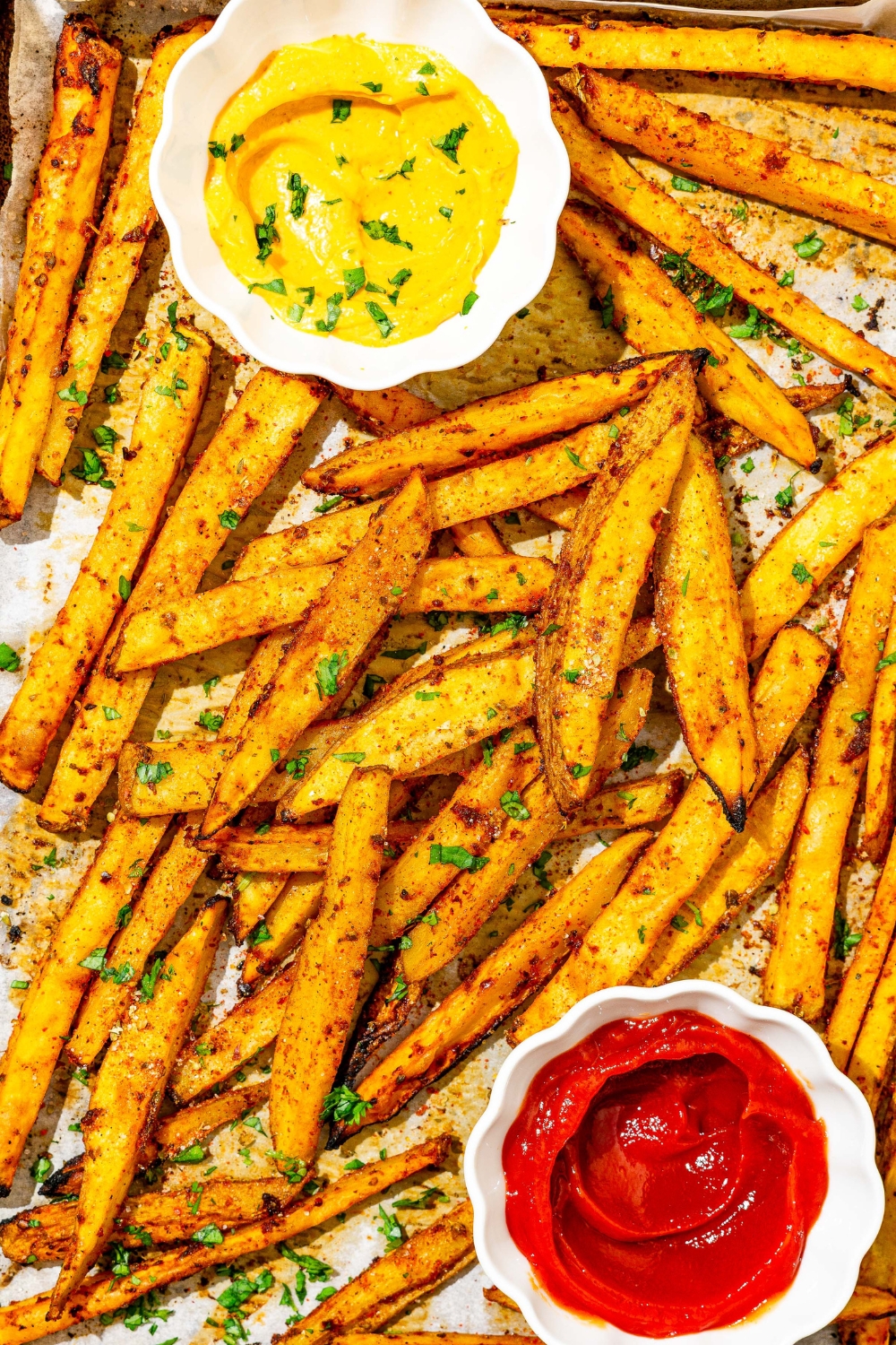 A baking sheet lined with parchment paper with cooked copycat Five Guys cajun fries. There is a small bowl of mustard and small bowl of ketchup on the baking sheet. Everything is garnished with fresh parsley.