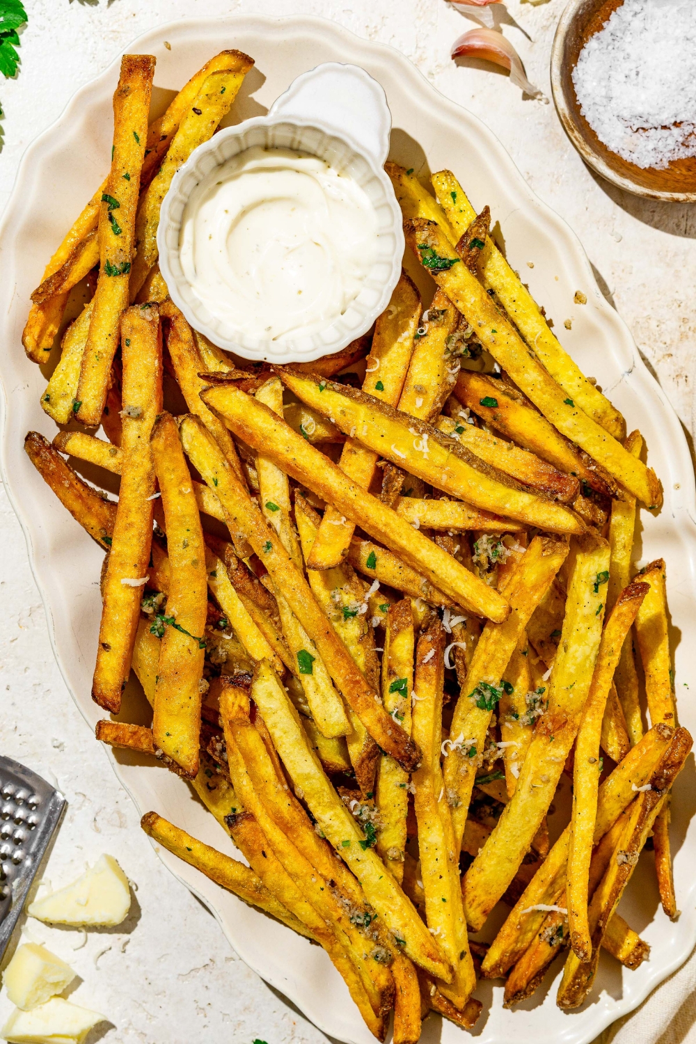 Garlic parmesan french fries on a white plate with a side of dipping sauce. The plate is on a white counter with a small side of salt.