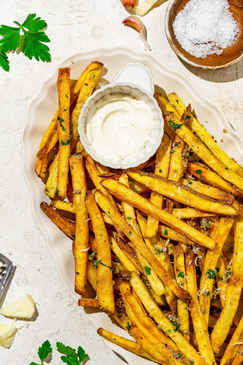Garlic parmesan french fries on a white plate with a side of dipping sauce. The plate is on a white counter with a small side of salt.