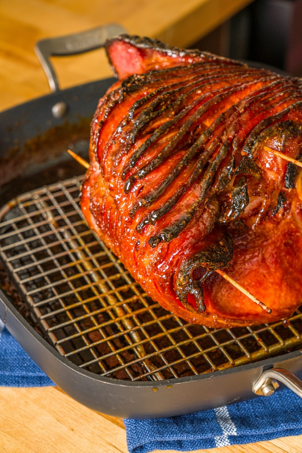 A whole glazed spiral ham on a roasting pan with a rack. The ham is sliced with toothpicks in the center. The pan is on a wooden counter.