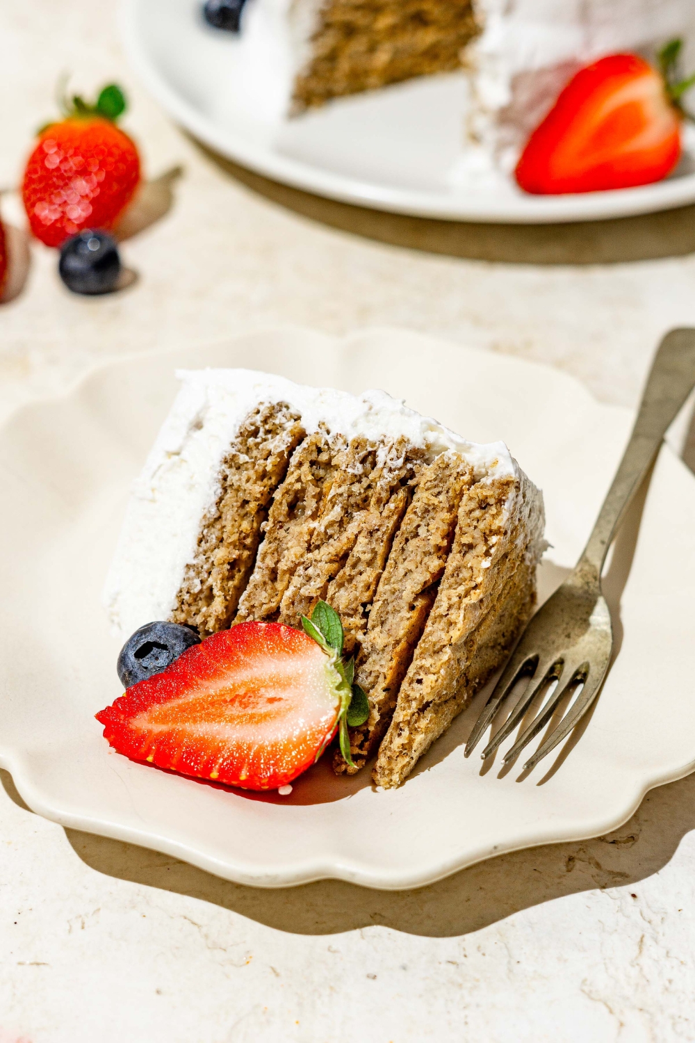 A slice of frosted banana smash cake on a white scalloped plate with a fork and sliced strawberry. The plate is on a tan counter with a whole banana smash cake.