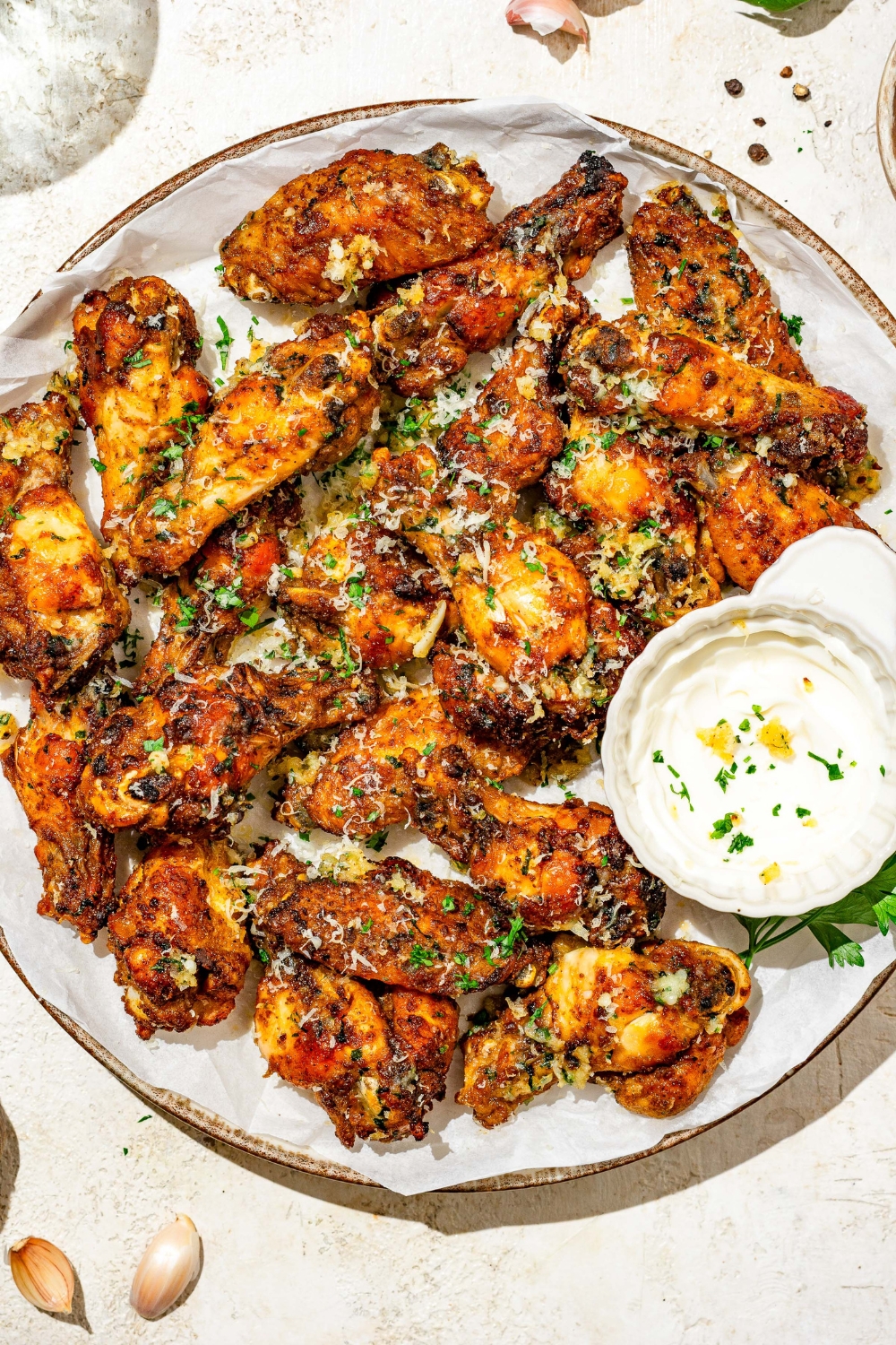 A plate of copycat Wingstop garlic parmesan chicken wings garnished with grated cheese and fresh parsley. There is a bowl of sauce on the plate. The plate is on a tan counter with small bowls of seasonings.