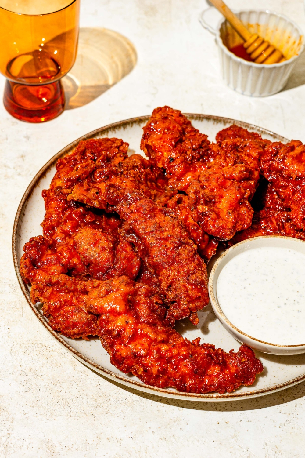 A plate of copycat Wingstop hot honey chicken tenders with a small bowl of ranch sauce. The plate is on a tan counter with a small bowl of honey with a honey stirrer.