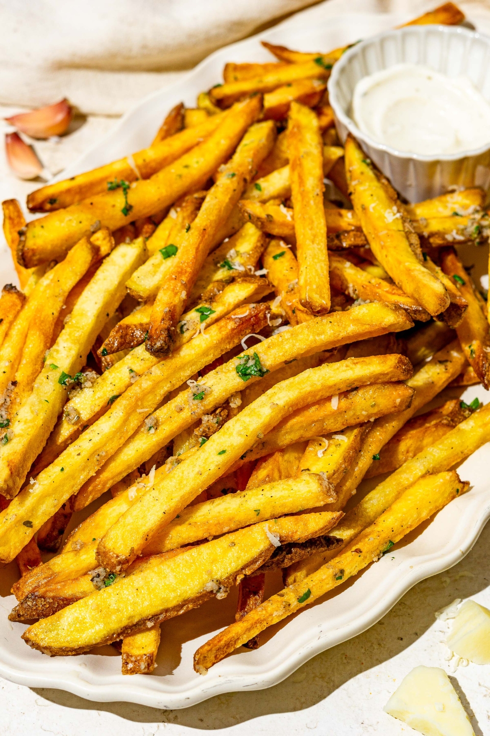 Garlic parmesan french fries on a white plate with a side of dipping sauce. The plate is on a white counter with a small side of salt.