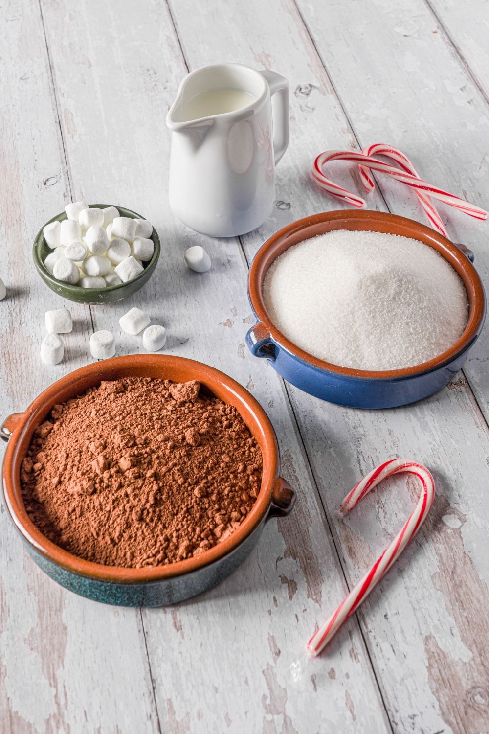 Bowls in various sizes containing ingredients to make hot cocoa mix including cocoa powder, sugar, a pitcher of milk, and small bowl of marshmallows.