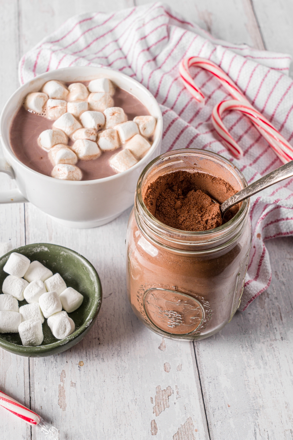 A mason jar with homemade hot cocoa mix with a spoon in the jar. The jar is on a wooden counter with a mug of hot cocoa topped with marshmallows, a small bowl of marshmallows, and candy canes.