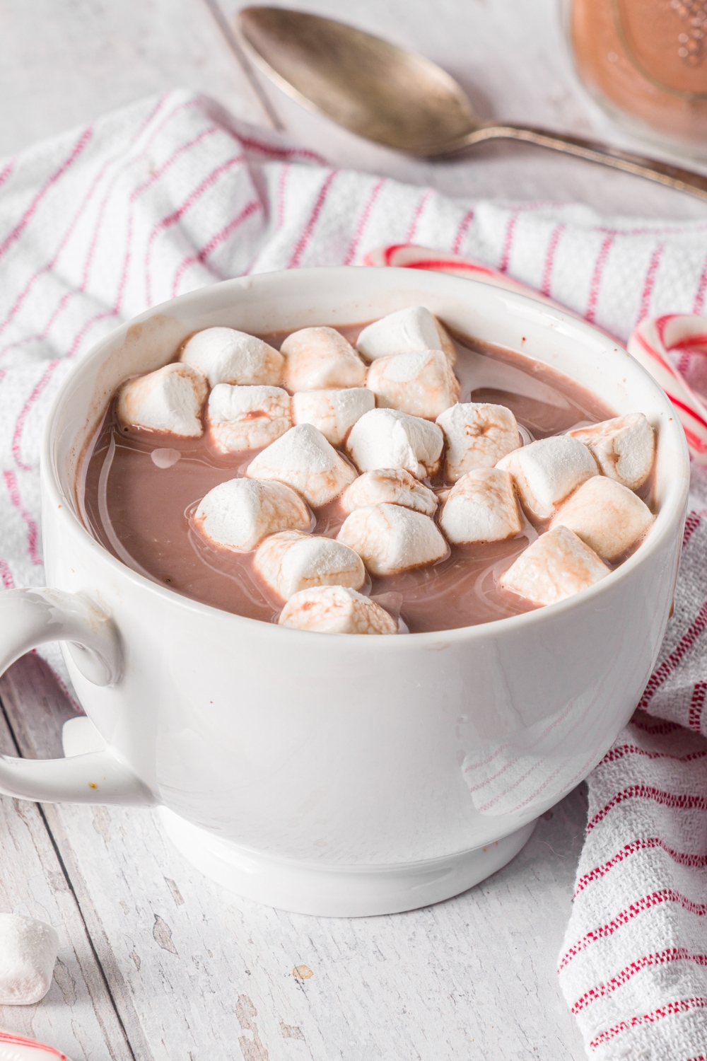 A white mug with homemade hot cocoa topped with marshmallows. The mug is on a wooden counter with a red striped napkin, spoon, and candy canes.