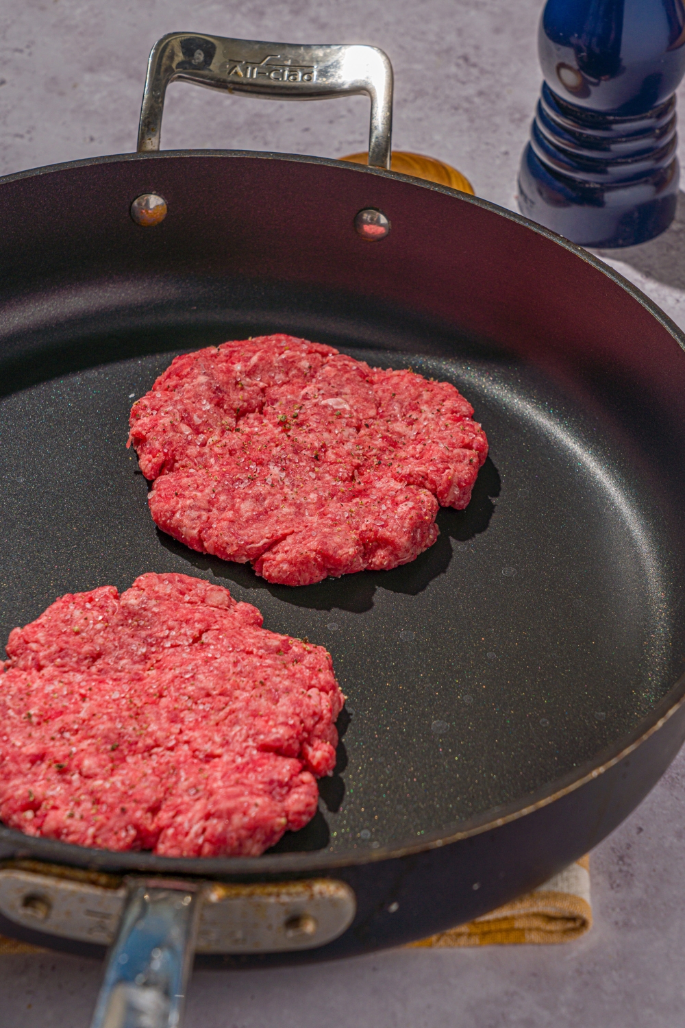 A skillet with two seasoned burger patties. The skillet is on a stone counter.