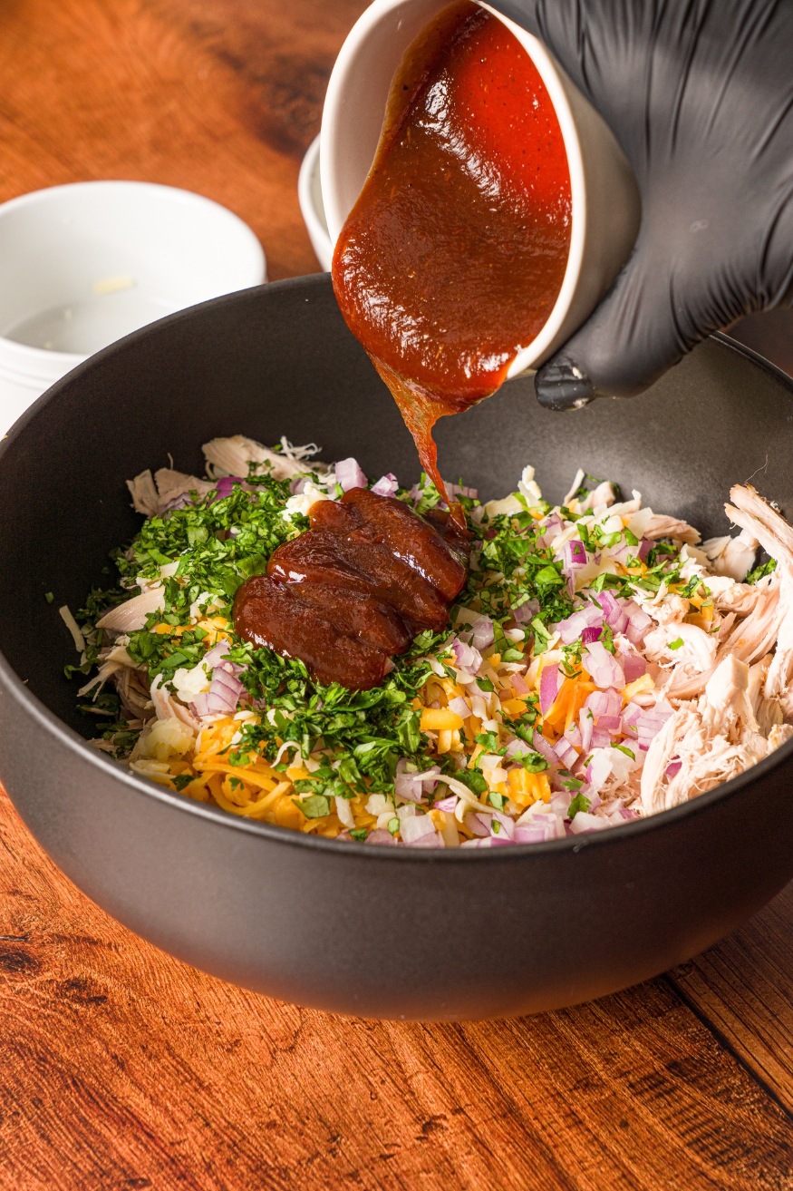 A black bowl with a shredded chicken mixture including diced onion, shredded cheese, and cilantro. A bowl of BBQ sauce is being poured into the mixture.