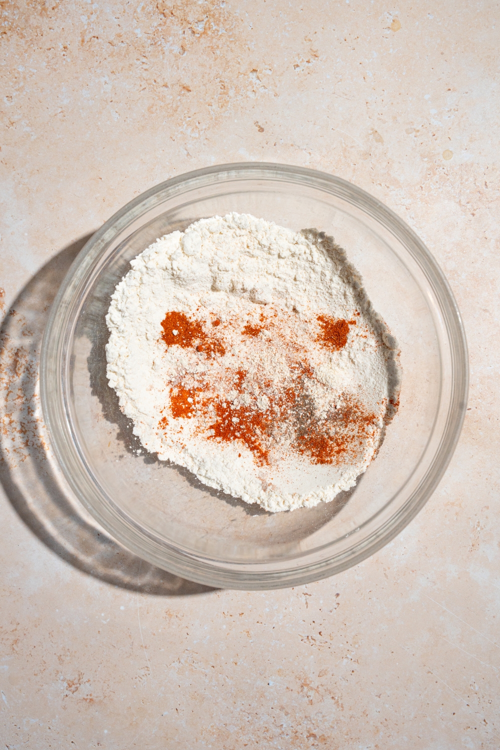 A glass bowl of seasonings including flour, cayenne pepper, salt, garlic powder, and more. The bowl is on a tan counter.