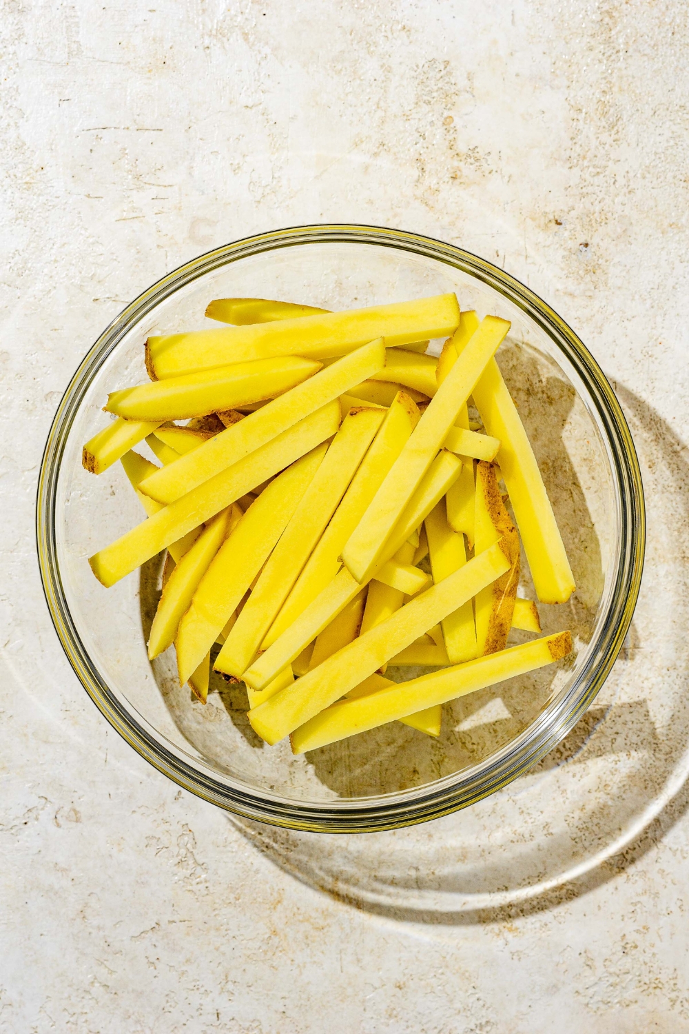 A glass bowl with sliced uncooked fries. The bowl is on a white counter.