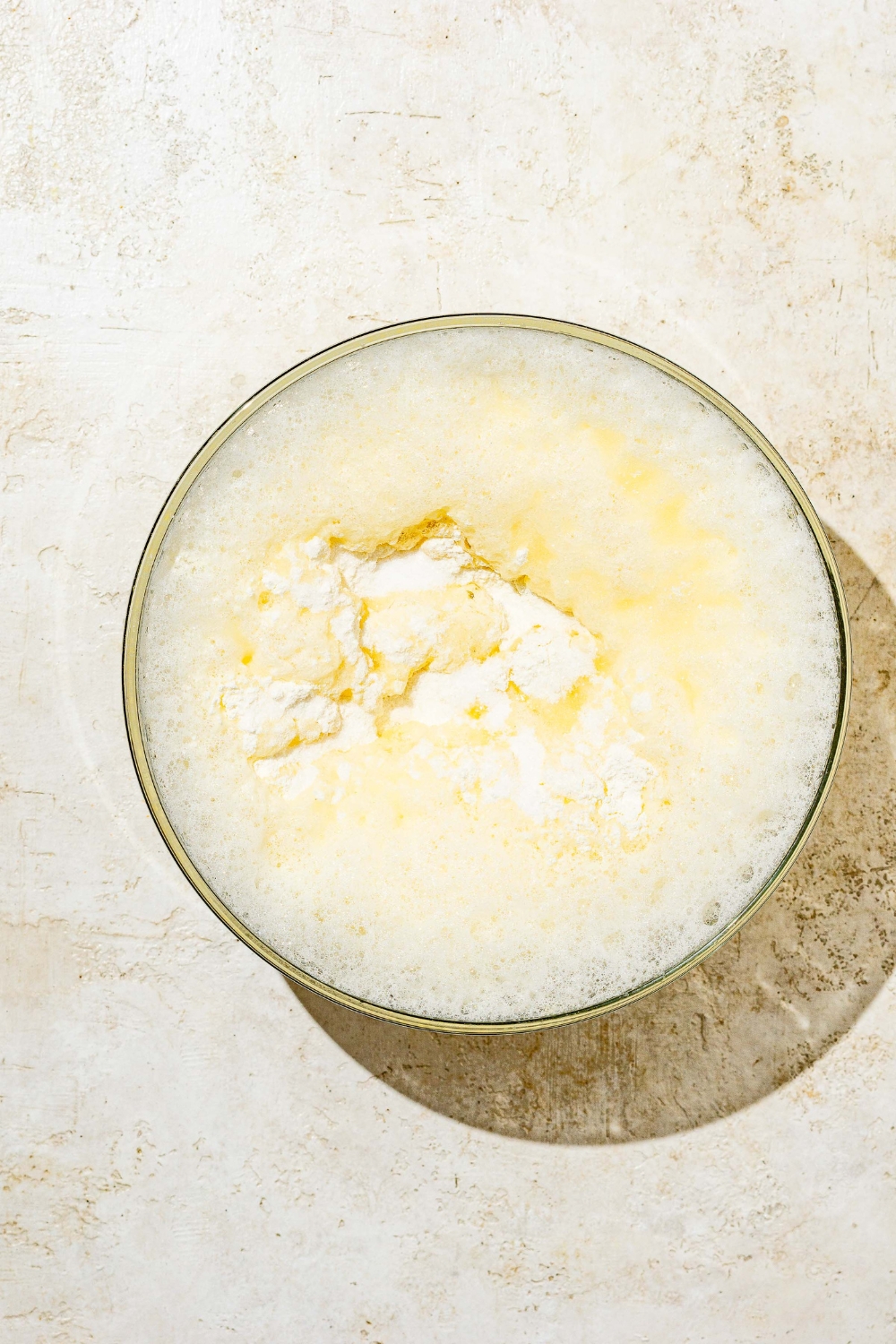 A glass bowl with blended ingredients to make Swedish pancake batter topped with flour. The bowl is on a white counter.