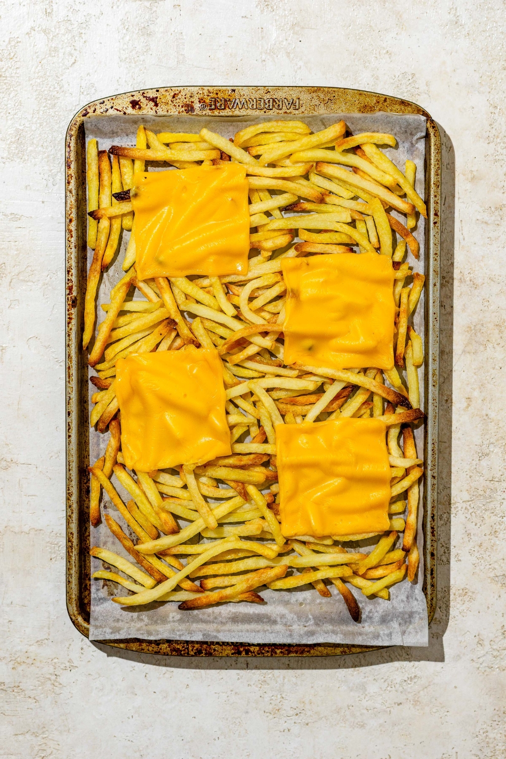 A baking sheet of baked fries topped with melted slices of American cheese. The sheet is on a tan counter.