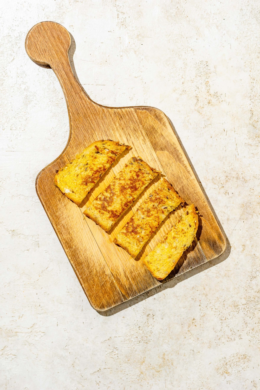 A wooden cutting board with a piece of baby french toast sliced into strips. The board is on a tan counter.