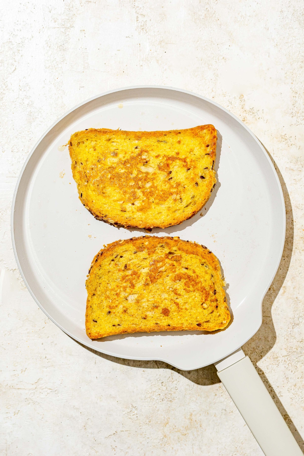 A ceramic skillet with two slices of baby french toast cooking. The skillet is on a tan counter.