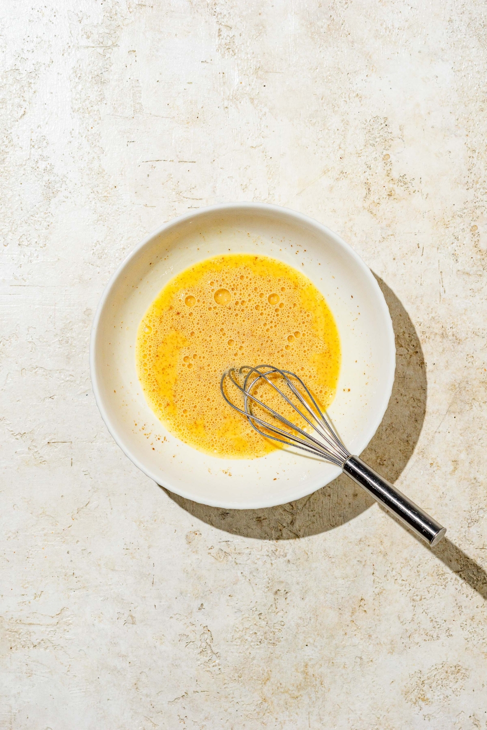 A small white bowl with a whisk mixing an egg mixture for french toast. The bowl is on a tan counter.