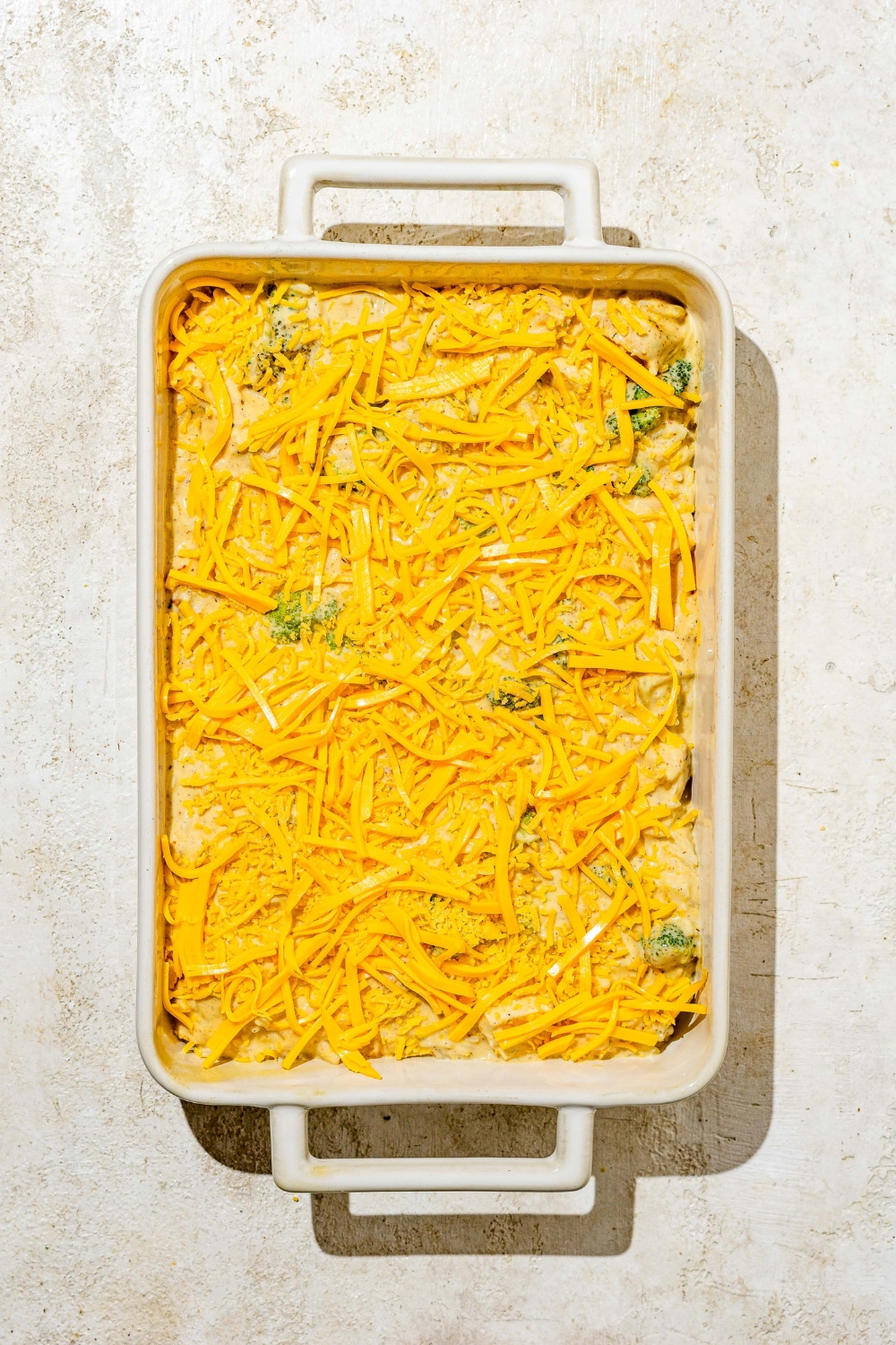 A baking dish with chicken broccoli and rice mixture topped with shredded cheddar cheese. The dish is on a tan counter.
