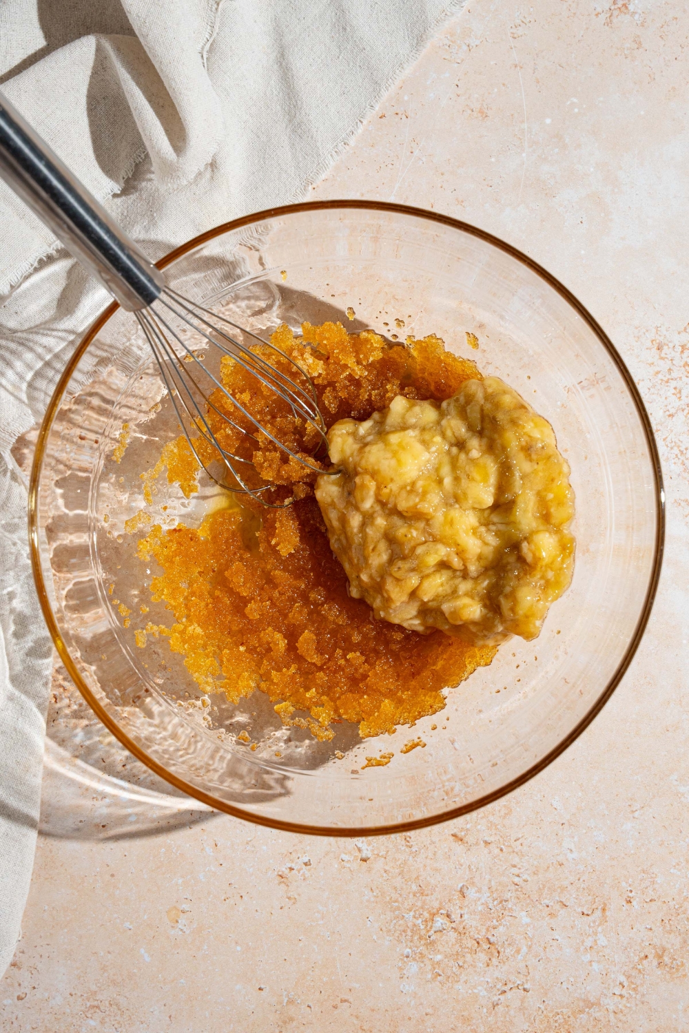 A glass bowl with a whisk mixing brown butter sugar mixture with mashed banana. The bowl is on a tan counter.