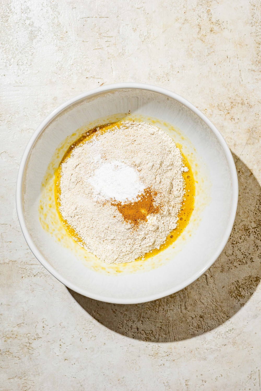 A white bowl with dry ingredients mixing with wet ingredients to make banana smash cake batter. The bowl is on a tan counter.
